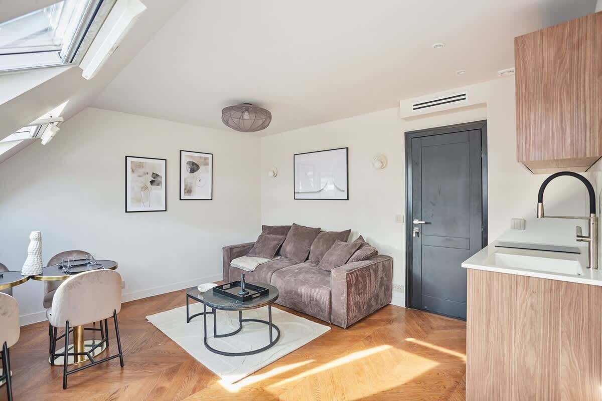 A modern apartment with a loft-style ceiling, a grey sofa, a dining area with a round table, and a kitchenette with a white countertop.