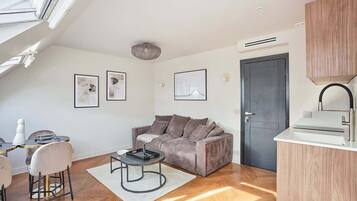 A modern apartment with a loft-style ceiling, a grey sofa, a dining area with a round table, and a kitchenette with a white countertop.