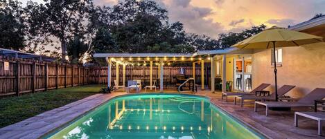 A backyard with a swimming pool, a covered patio area with bookshelves, and a fenced yard.