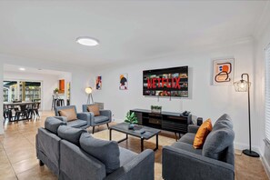 A modern living room with grey sofas, a coffee table, and a TV displaying 'NETFLIX'. There is a dining area in the background with a table and chairs.