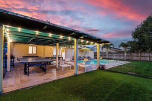 Uma área externa coberta com mesa de pingue-pongue, cadeiras e piscina.