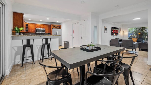A modern kitchen with wooden cabinets, a stainless steel microwave, and a refrigerator. There is a dining area with a large table and chairs, and a living room with a sofa and a coffee table. - Fort Lauderdale Oasis: 4BR, Pool & Game Room (Fort Lauderdale)
