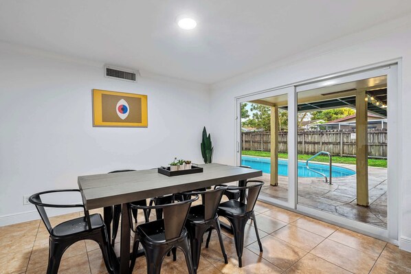 A dining area with a table and chairs, a framed picture on the wall, and a view of a backyard with a pool.