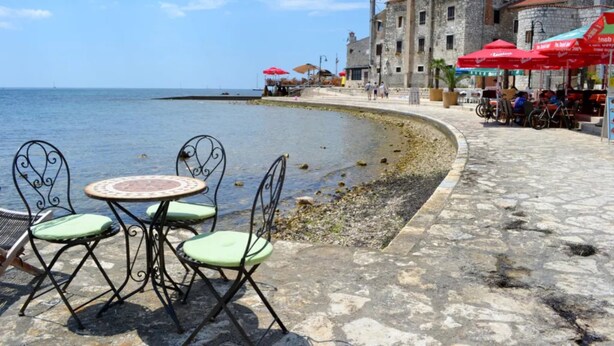 A coastal area with stone tables and chairs, a stone building, and red umbrellas. - Charming Apartment in Umag With Balcony (Umag)