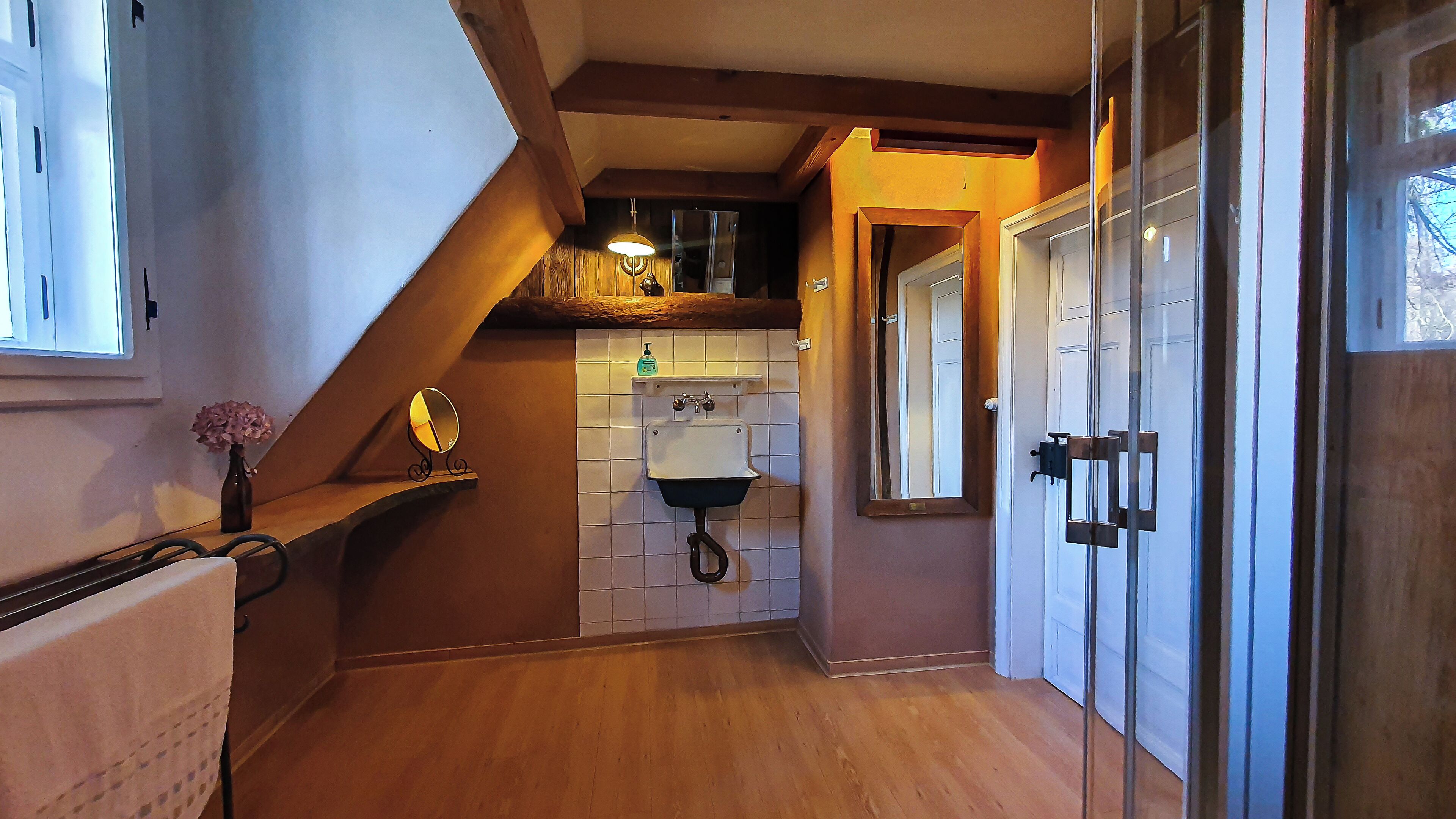 A narrow hallway with a wooden floor, a white tiled wall, a small sink with a soap dispenser, and a mirror on a shelf.