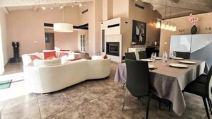 Een ruime woonkamer met een gebogen witte bank, een eettafel voor vier personen en een keuken op de achtergrond.