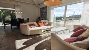 Een ruime woonkamer met een groot raam, een witte bank, een salontafel en een eethoek met stoelen en een tafel.