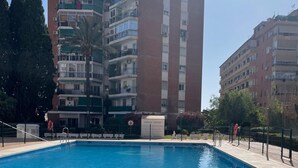 A rectangular swimming pool with a tiled edge, surrounded by a low fence. In the background, there are multi-story residential buildings with balconies and windows.