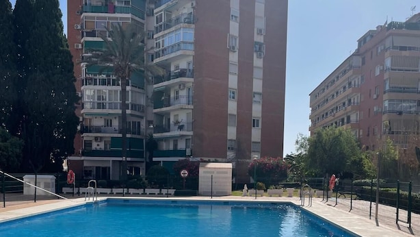Una piscina rectangular con borde de azulejos, rodeada por una valla baja. Al fondo, edificios residenciales de varios pisos con balcones y ventanas.