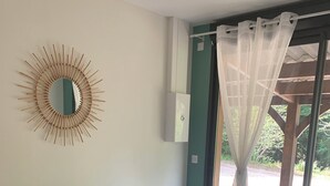 Ein Schlafzimmer mit einem Bett, einem Wandspiegel, einem Fenster mit durchsichtigen Vorhängen und Blick ins Grüne.