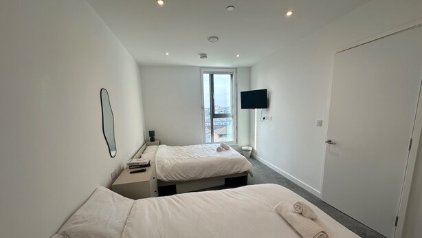 A modern hotel room with two beds, a flat-screen TV mounted on the wall, and a large window offering a city view. - 2 Bedroom Apartment - Sleeps 6 (Manchester)