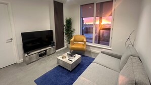 A modern living room with a flat-screen TV, a sofa, a coffee table, and a view of a sunset through large windows. - 2 Bedroom Apartment - Sleeps 6 (Manchester)