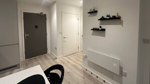 A modern living room with a wooden floor, a black chair, a white sofa, and a white wall with black shelves holding decorative items. - 2 Bedroom Apartment - Sleeps 6 (Manchester)