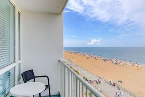 A balcony with a white table and chair, overlooking a beach with people and umbrellas. - Boardwalk Resort & Villas - 1BR/1BA - Oceanfront! (Virginia Beach)