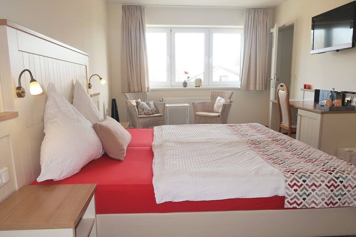 Ein Schlafzimmer mit Bett, Nachttischen, einem Sessel, einem Schreibtisch, einem Fernseher und einem Fenster mit Vorhängen.