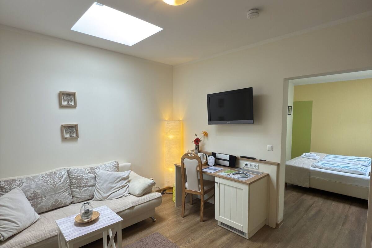 Ein kompakter Wohnraum mit Sofa, kleinem Tisch, Schreibtisch mit Stuhl, Bett, Fernseher und Dachfenster.