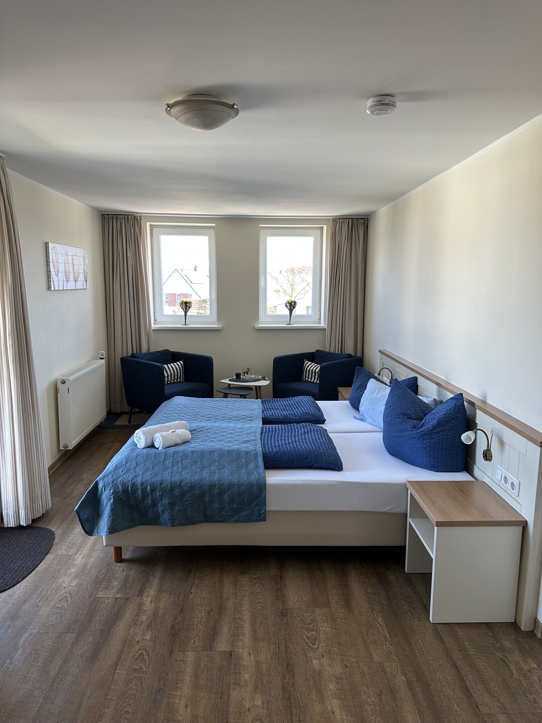 A modern hotel room with a large bed, two bedside tables, a small desk, and two windows with curtains.