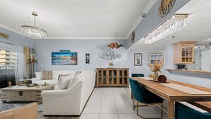Ein modernes Wohnzimmer mit einem weißen Sofa, einem Holzesstisch und smaragdgrünen Stühlen. Es gibt einen Holzschrank mit Glastüren und eine Kronleuchter.