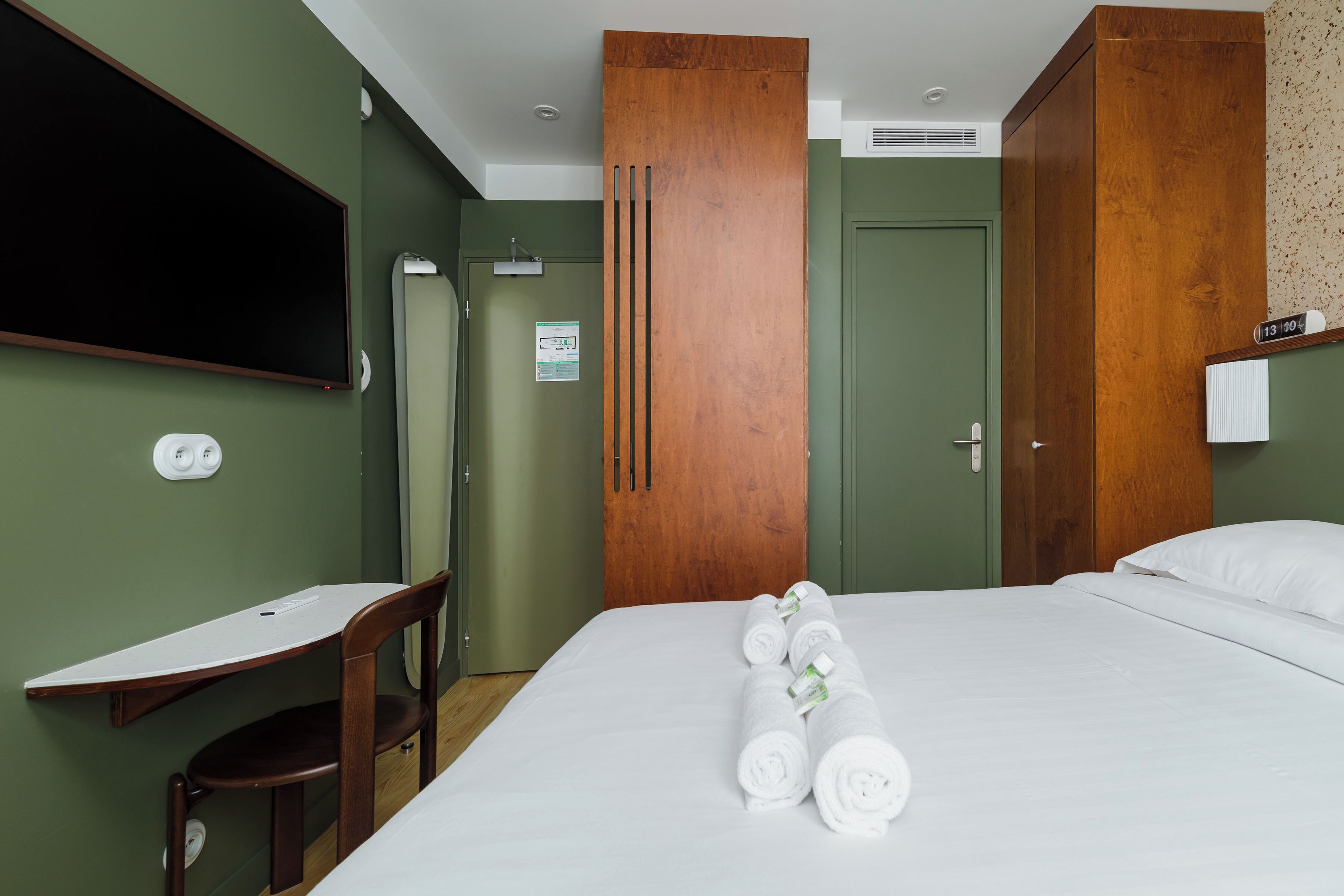 A hotel room with a bed, a desk, a chair, and a wooden wardrobe.