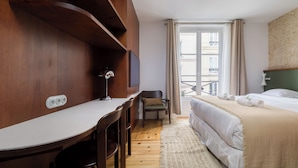 Ein Hotelzimmer mit Holzschreibtisch, einem Stuhl, einem Bett mit beiger Decke, einem Fernseher an der Wand und einem Fenster mit Vorhängen.