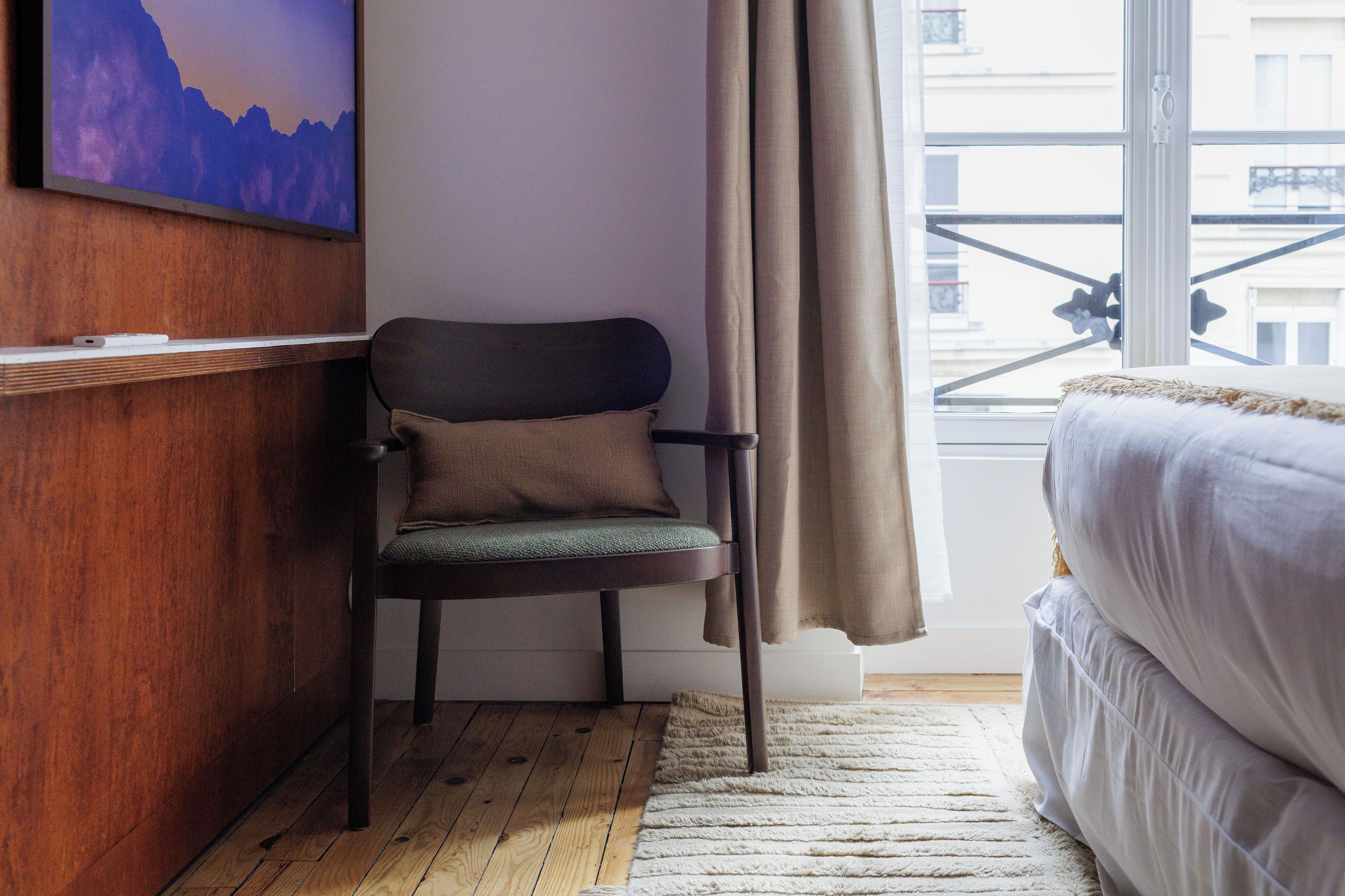 Ein Zimmer mit einem Holztisch, einem Stuhl, einem Bett und einem Fenster mit Vorhängen, mit Blick auf Gebäude.
