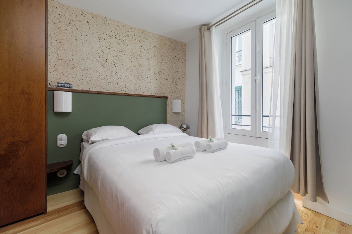 Habitación de hotel con una cama grande, cabecera verde, ventana con cortinas y una lámpara fijada a la pared.