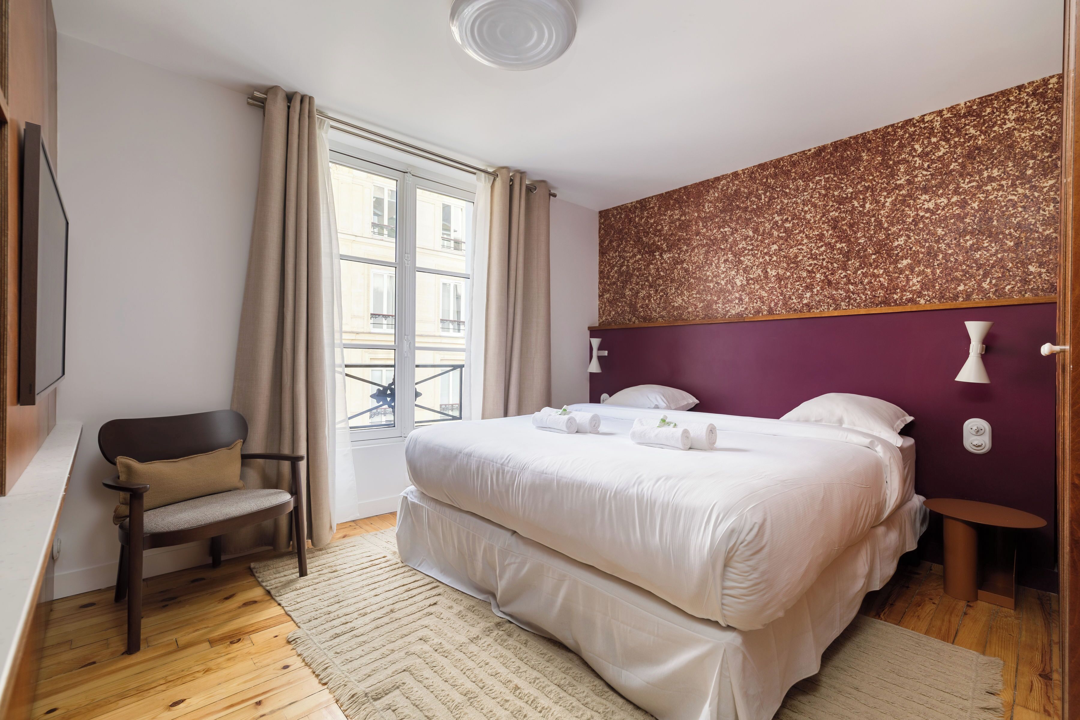 A bedroom with a large bed, a chair, a window with curtains, and a wall with a textured pattern.