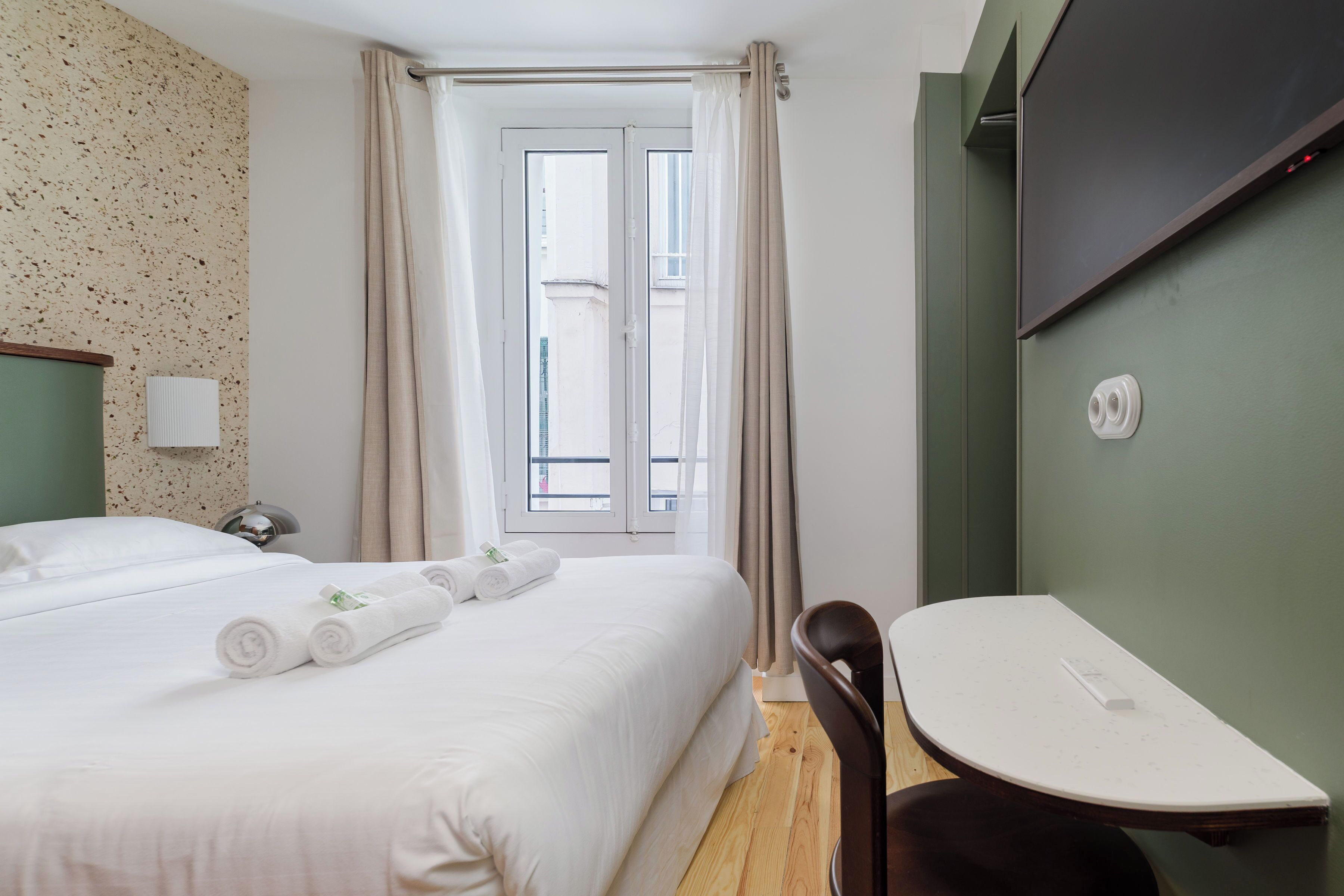 Ein Hotelzimmer mit Bett, Schreibtisch, Stuhl, einem Fenster mit Vorhängen und einem an der Wand befestigten Fernseher.