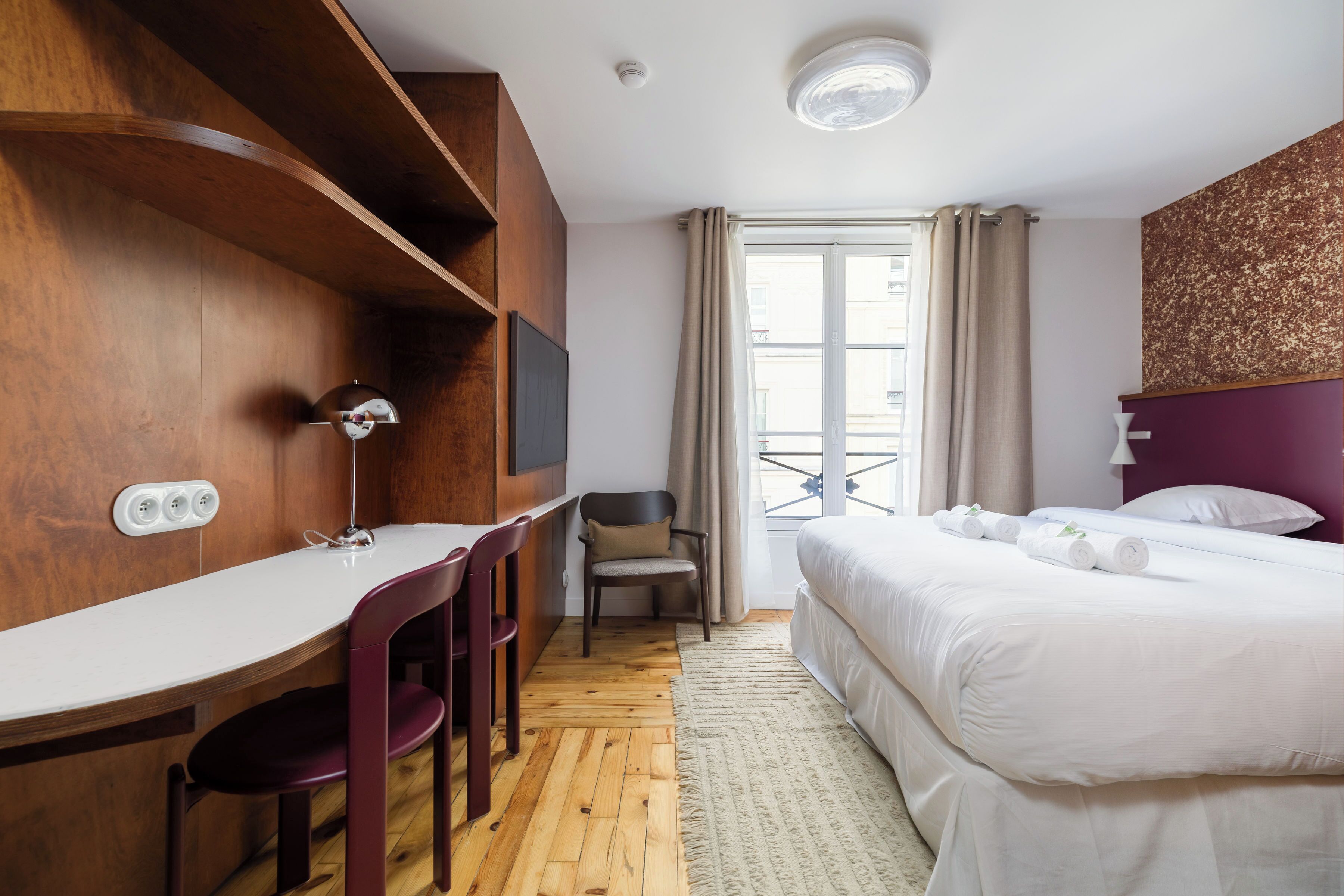 Une chambre d’hôtel avec un bureau en bois, un lit, une chaise et une fenêtre avec des rideaux.