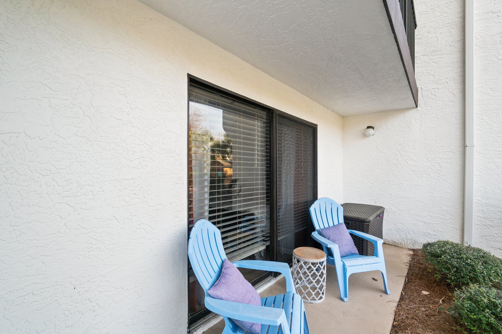 Un espace patio avec deux chaises bleues et une petite table ronde, une fenêtre avec des stores et un petit arbuste.