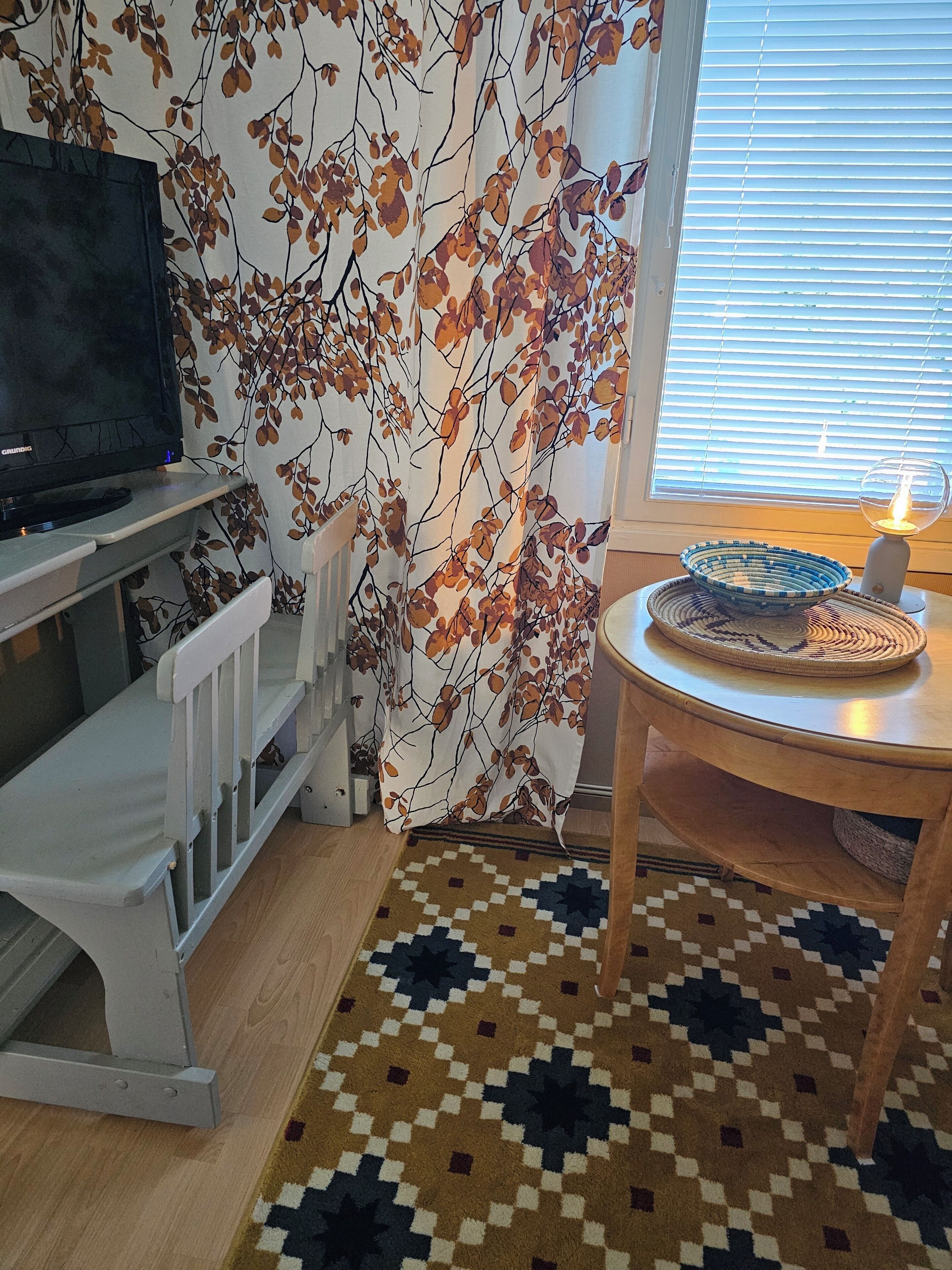 Une pièce avec un téléviseur posé sur un meuble, un rideau à motifs, une table avec une bougie et un tapis à motifs.