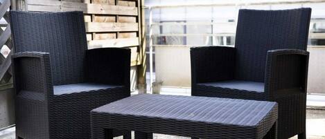 Two black wicker chairs and a matching table on an outdoor balcony with a wooden fence and a glass building in the background.