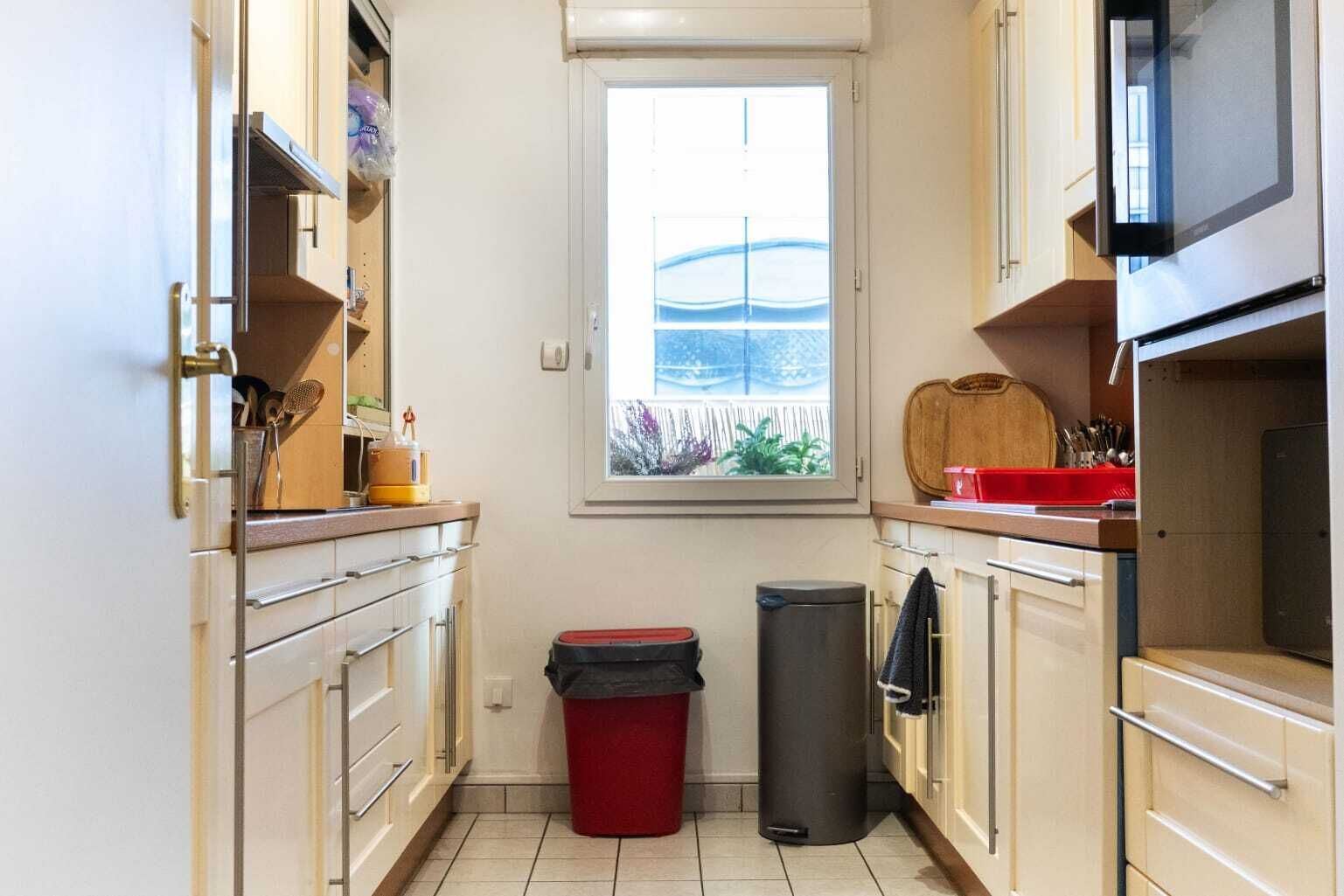 Une cuisine étroite avec des meubles blancs, une fenêtre donnant sur un beau paysage, un four à micro-ondes et une poubelle.
