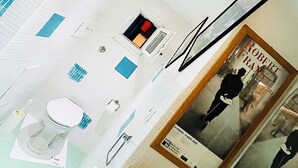 A modern bathroom with a white sink, toilet, and shower area. There is a framed picture of a person on the wall and a colorful abstract painting above the sink. - Hackert Art+Room (Caserta)