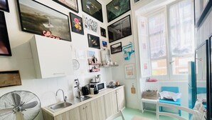 A kitchen with white cabinets, a sink, a microwave, and a fan. There are framed pictures on the wall and a window with a view outside. - Hackert Art+Room (Caserta)