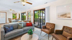 A spacious living room with a grey sofa, a brown armchair, and a coffee table. There is a ceiling fan, a dining area with a table and chairs, and large windows offering a view of the outdoors. - Sunset Escape - Fit For 8! (Gravois Mills)