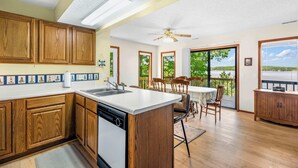 A spacious kitchen with wooden cabinets, a dishwasher, and a large island. There is a dining area with a table and chairs, and a view of the outdoors through large windows. - Sunset Escape - Fit For 8! (Gravois Mills)