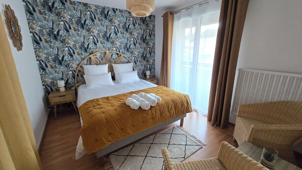 Un lit bien fait, avec une couette de couleur moutarde, une tête de lit en osier et un papier peint orné de motifs de feuilles tropicales.