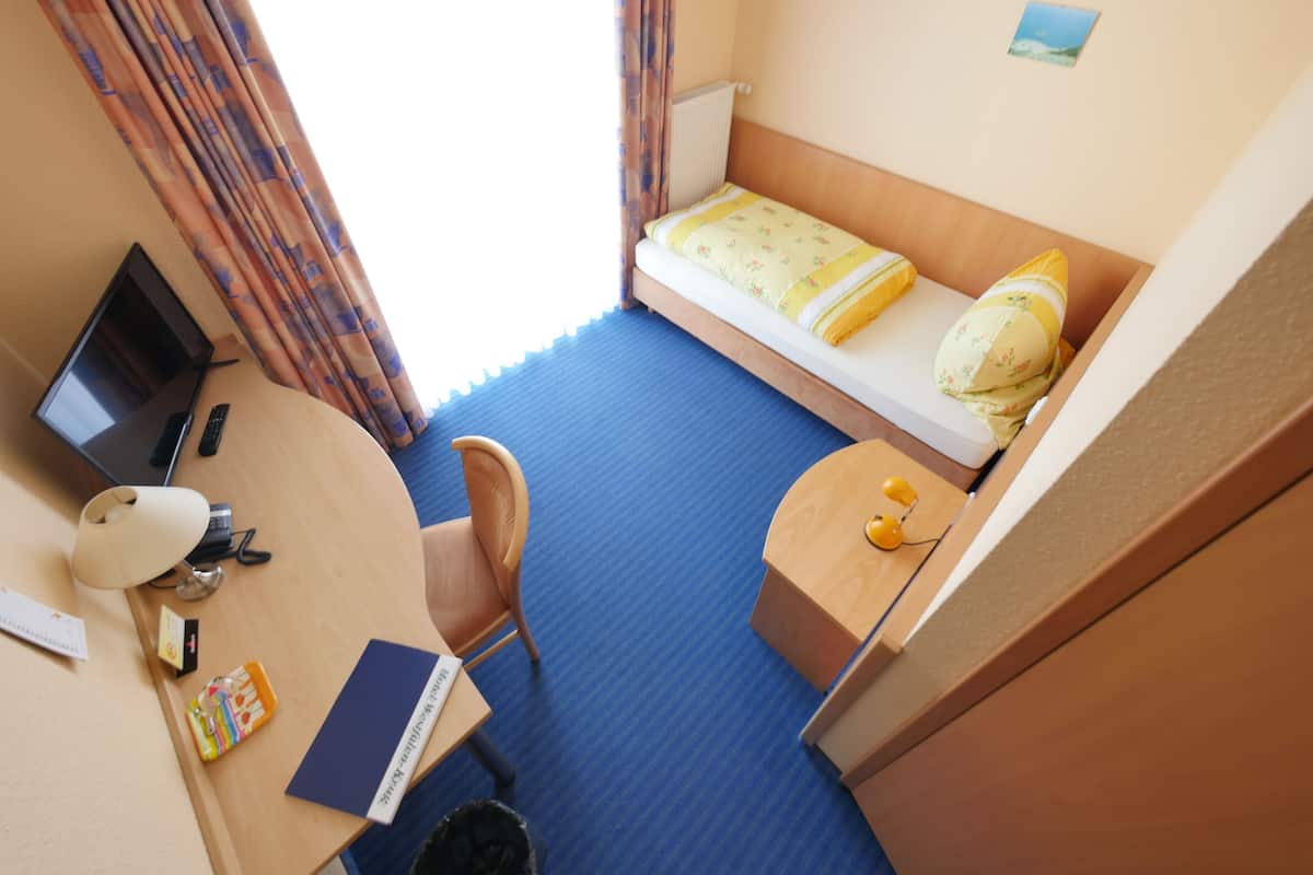 Een kleine hotelkamer met een enkel bed, een bureau met een computer, een stoel en een bijzettafeltje.