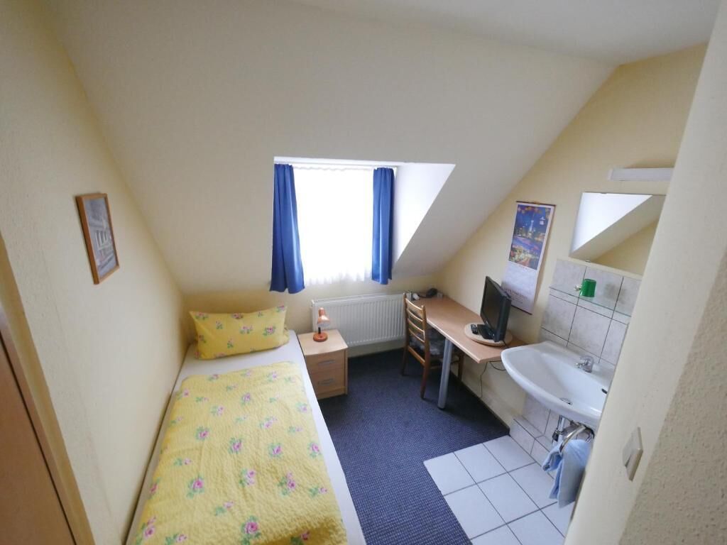 Una habitación pequeña y bien iluminada, con cama, escritorio, computadora y baño.