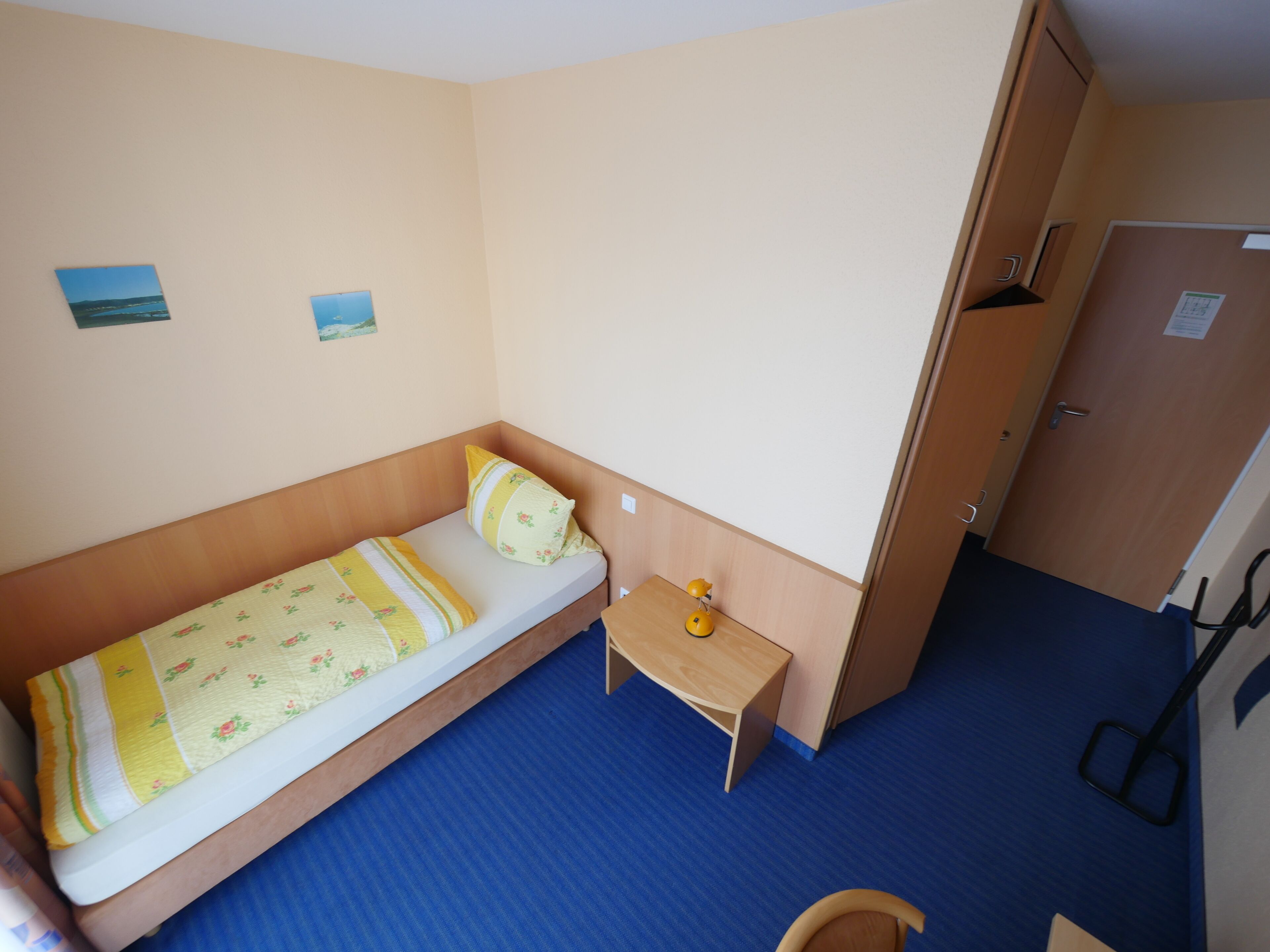 A single-bed room with a wooden headboard, a small bedside table with a lamp, and a door to the right.