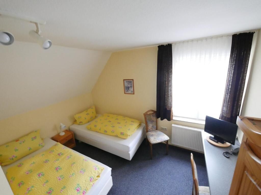 Una habitación de hotel pequeña y sencilla, con una cama, una silla, un escritorio con ordenador y una ventana con cortinas.