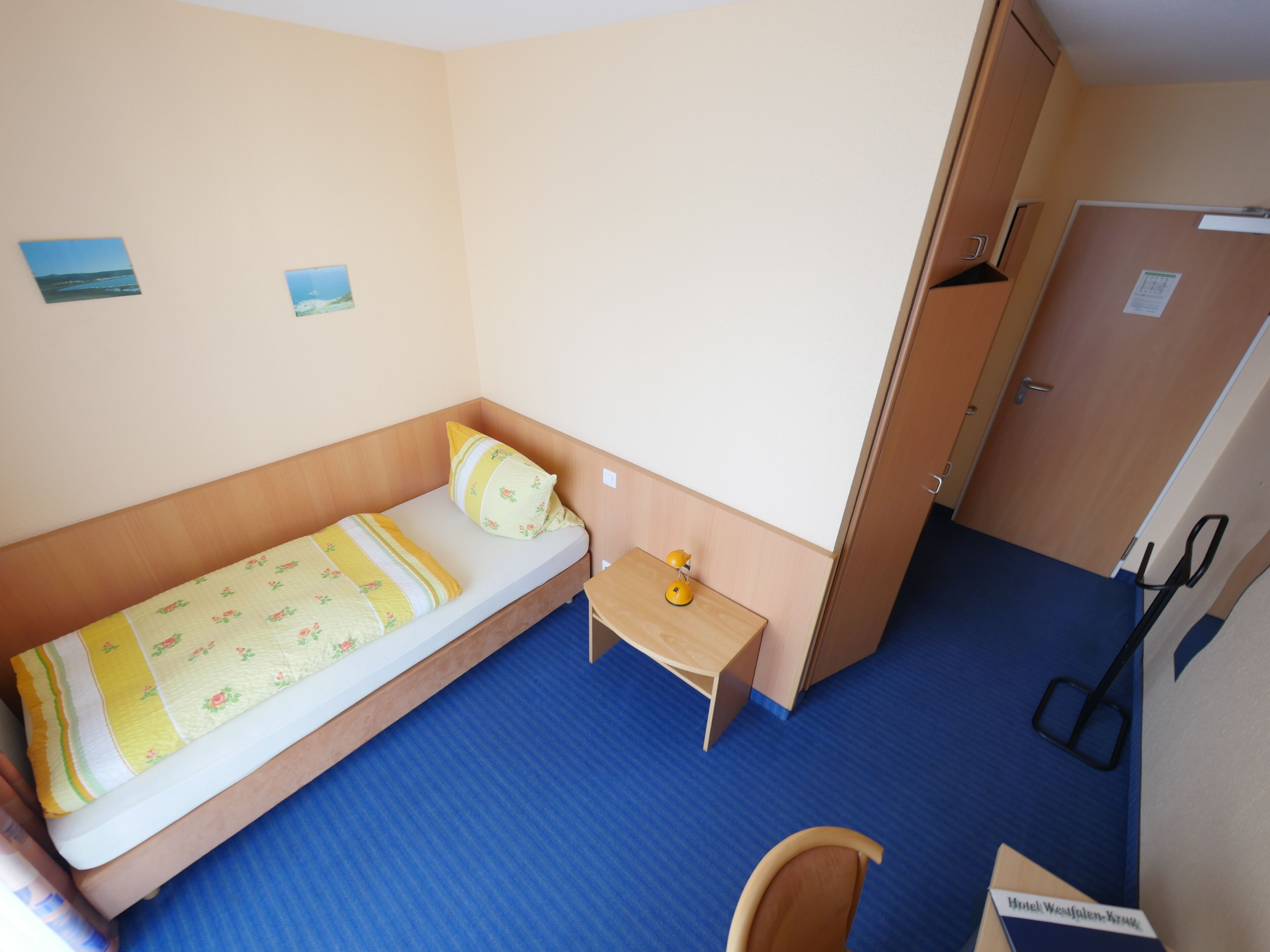 Um quarto pequeno com uma cama de solteiro, cabeceira de madeira, mesa de cabeceira com abajur, pequena escrivaninha e porta que dá para outro cômodo.