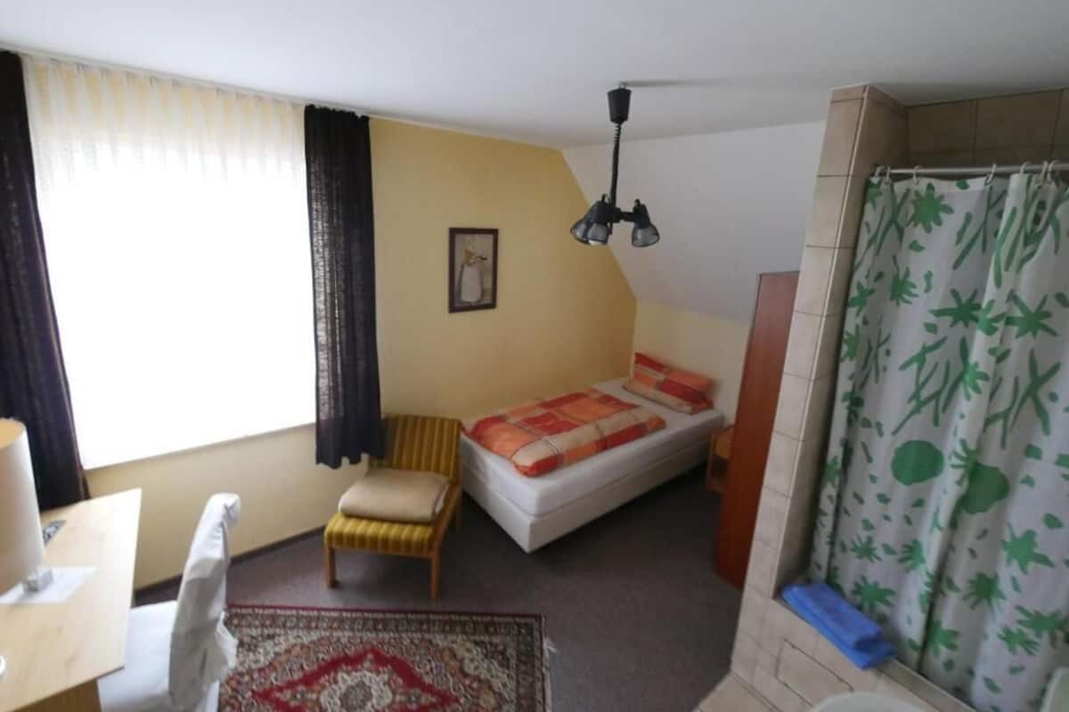 Een kleine, compacte kamer met een bed, een stoel, een bureau en een douche.