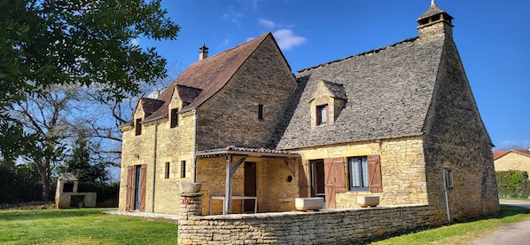 Une maison en pierre avec un toit à forte pente, une cheminée et un petit porche agrémenté d’une table et de chaises.