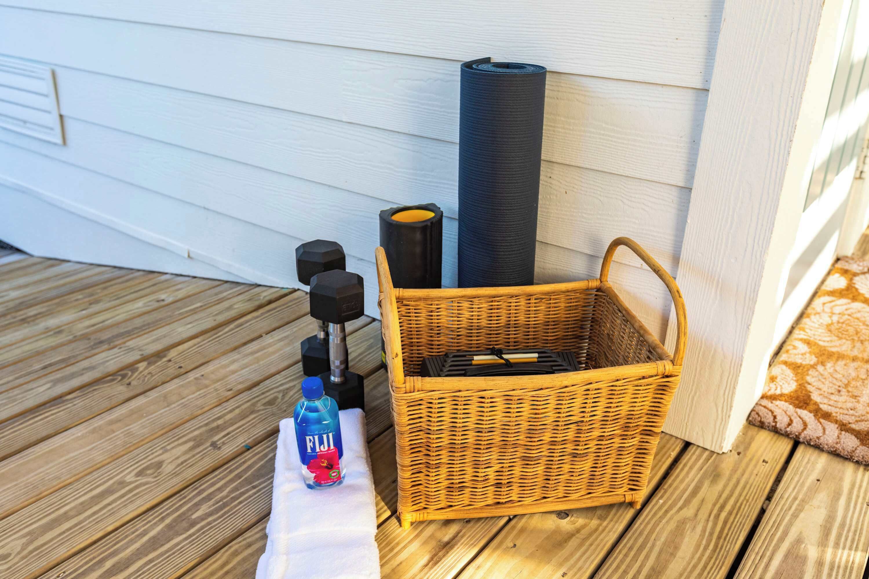 A wooden deck with a white wall, a wicker basket, a black cylindrical object, and a bottle of Fiji water.