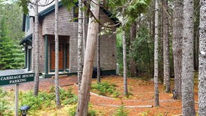 Une cabane dotée d’un porche, entourée de grands arbres et agrémentée d’un panneau indiquant l’emplacement du parking pour les voitures.