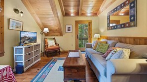 Un salon chaleureux avec un plafond en bois, un canapé, une table basse, un téléviseur, une bibliothèque et une fenêtre.