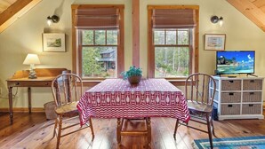 Un coin repas comprenant une table, des chaises, une lampe, un téléviseur et une vue sur les arbres à travers les fenêtres.