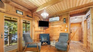 Interior de una cabaña de madera con dos sillones, una chimenea, un televisor y vistas a través de un ventanal.
